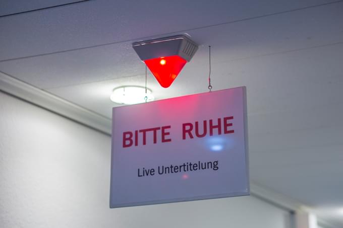 Rote Signallampe im Sendestudio, darunter ein Schild mit dem Text "Bitte Ruhe, Live Untertitelung"
